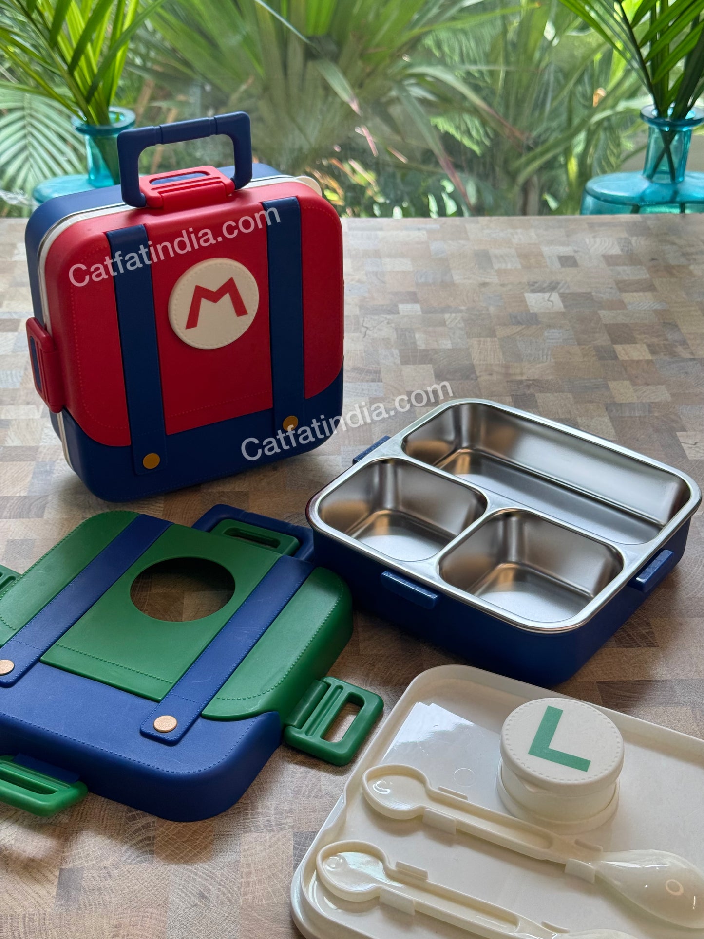 A children's lunch box set displayed on a wooden table, featuring a red and blue lunchbox with  a stainless steel three-compartment tray, and a cream-colored plastic spoon and cup set. A 'Catfatindia.