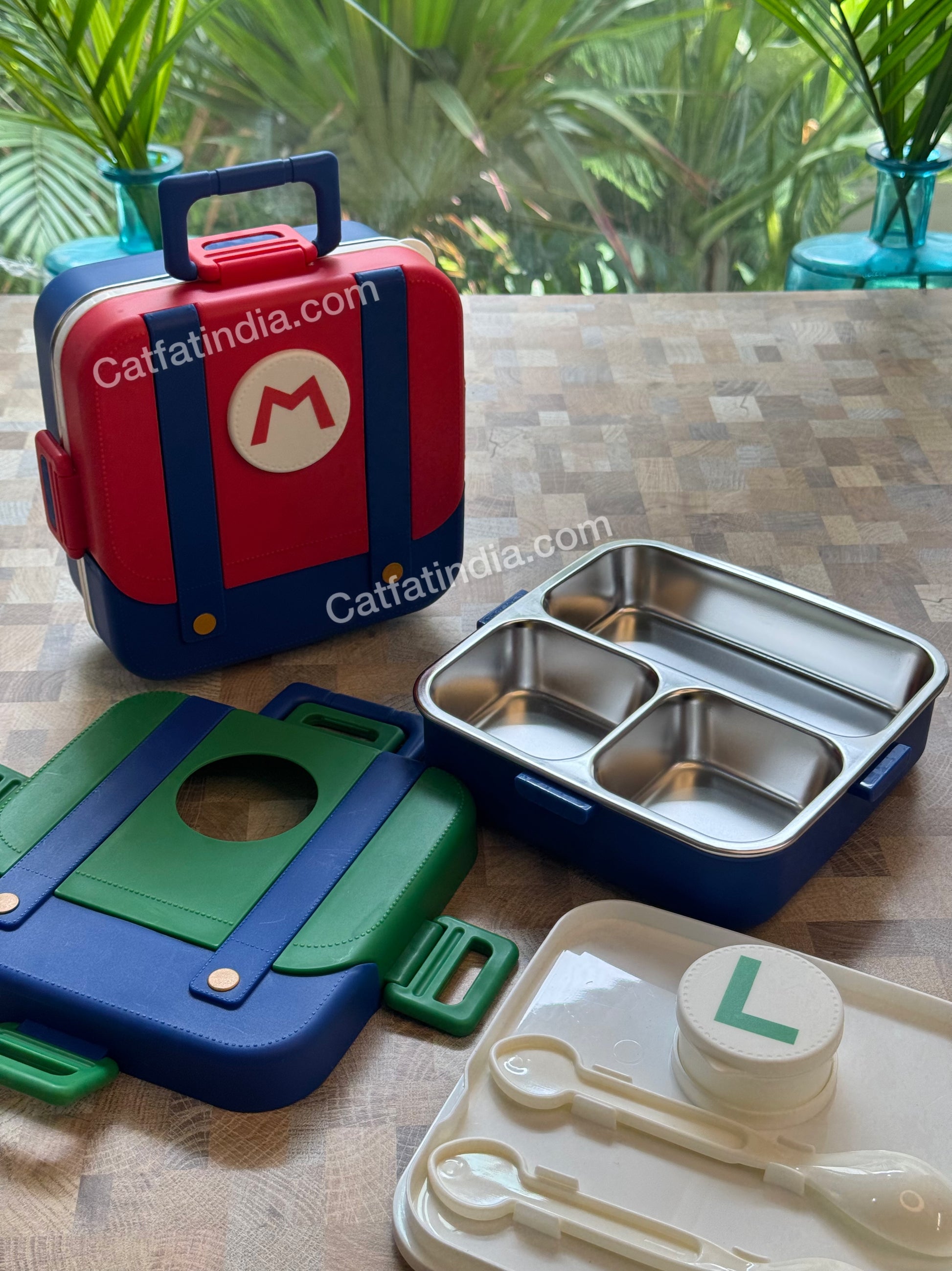 A children's lunch box set displayed on a wooden table, featuring a red and blue lunchbox with  a stainless steel three-compartment tray, and a cream-colored plastic spoon and cup set. A 'Catfatindia.